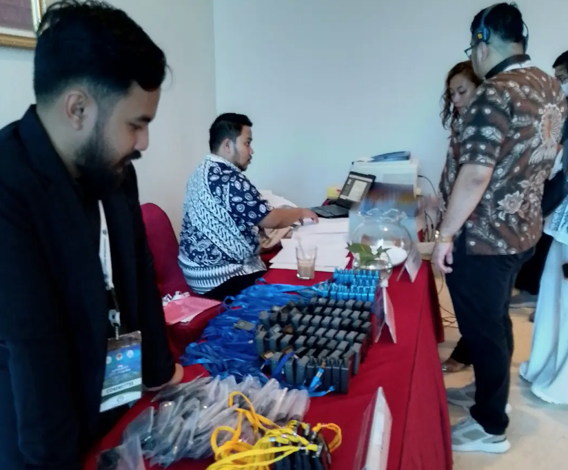 Meja registrasi event dengan receiver TGS/panaguide berjajar lengkap dengan lanyard, dibagikan ke peserta yang memakai headset]